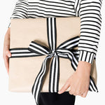 Woman in a black-and-white striped top holding a kraft-paper-wrapped box tied with striped ribbon. Gift wrapping and ribbon are not included. | Women's Mystery Clothing Box | Lacson Ravello